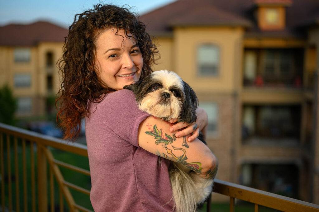 Woman with curly hair in a purple shirt stands on a balcony, smiling and holding a small black-and-white dog. Residential buildings are visible in the background.