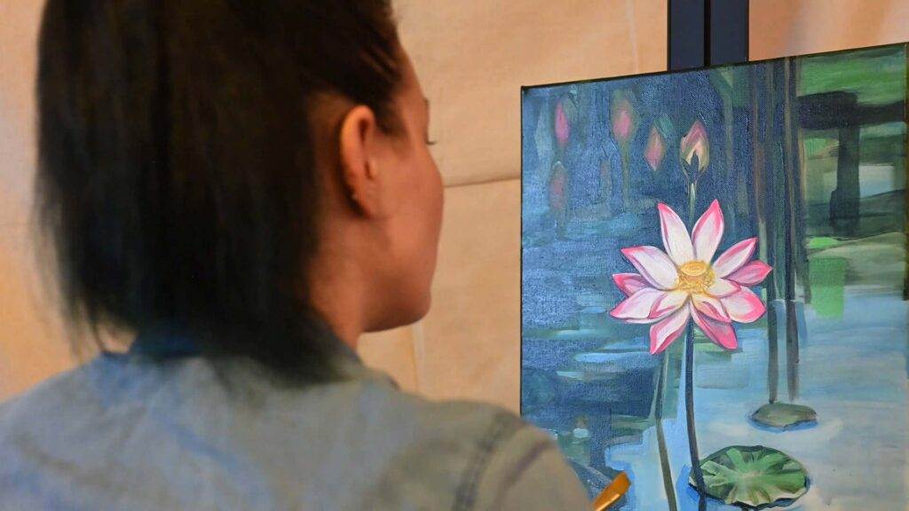 A person is painting a detailed image of a pink and white flower on a blue and green background. The person’s face is not visible as they work on the canvas.