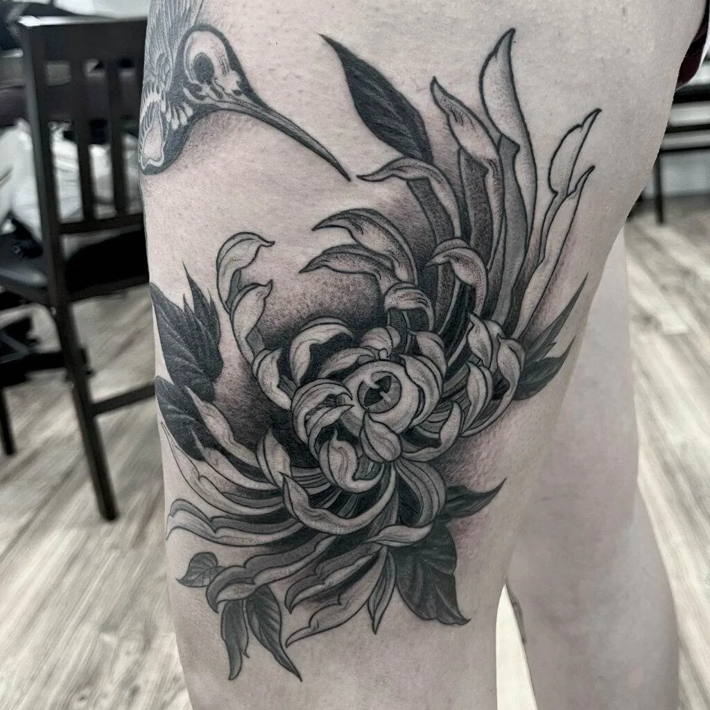 A grayscale tattoo on a person's thigh depicts an intricate chrysanthemum flower and a hummingbird.