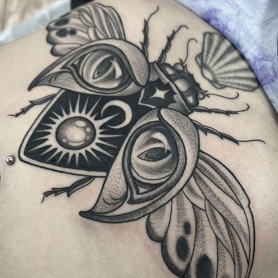 A detailed black and gray tattoo of an abstract moth with eyes on its wings and geometric shapes on an area of skin.