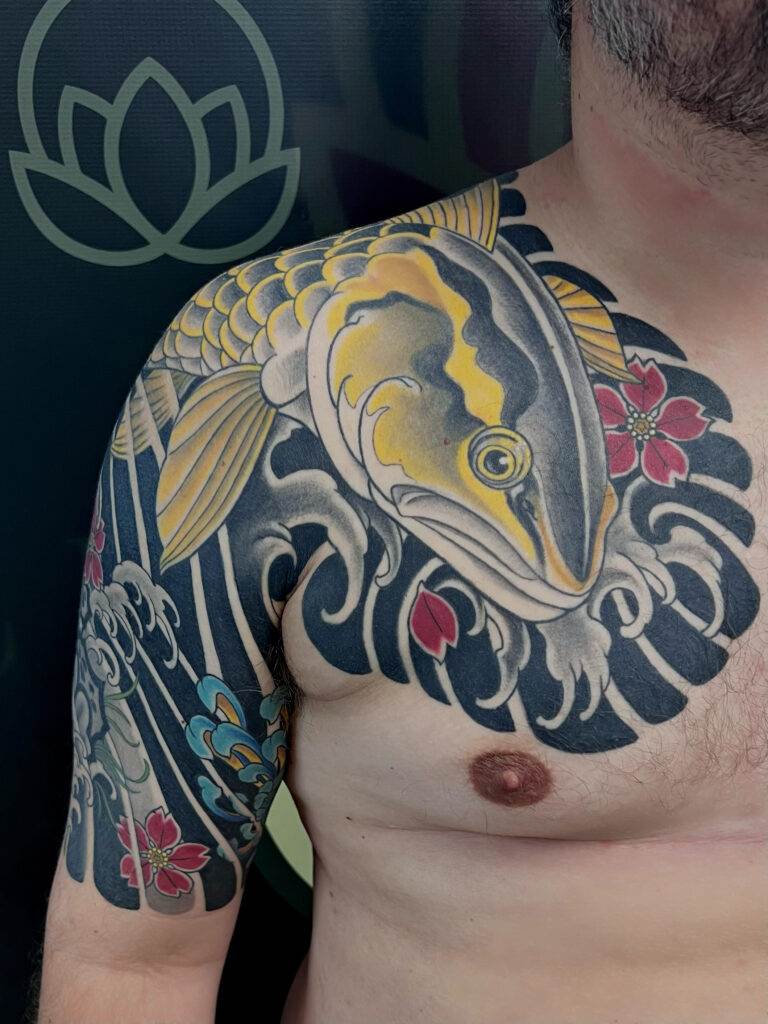 A close-up of a man's chest and shoulder showcasing a detailed tattoo of a koi fish surrounded by waves and flowers.