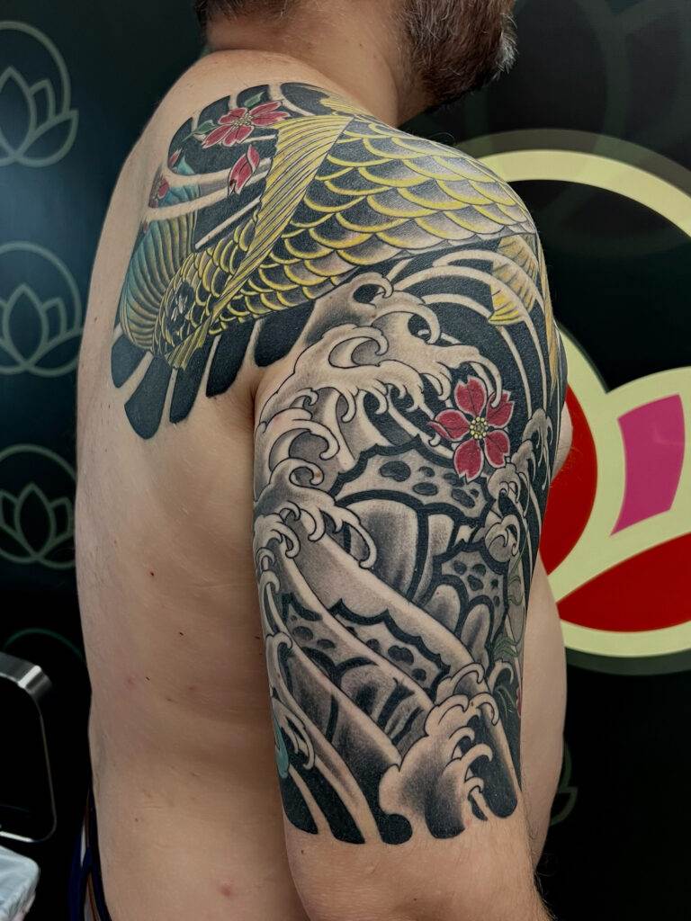 A person with an intricate tattoo on their arm featuring a koi fish, waves, and cherry blossoms.