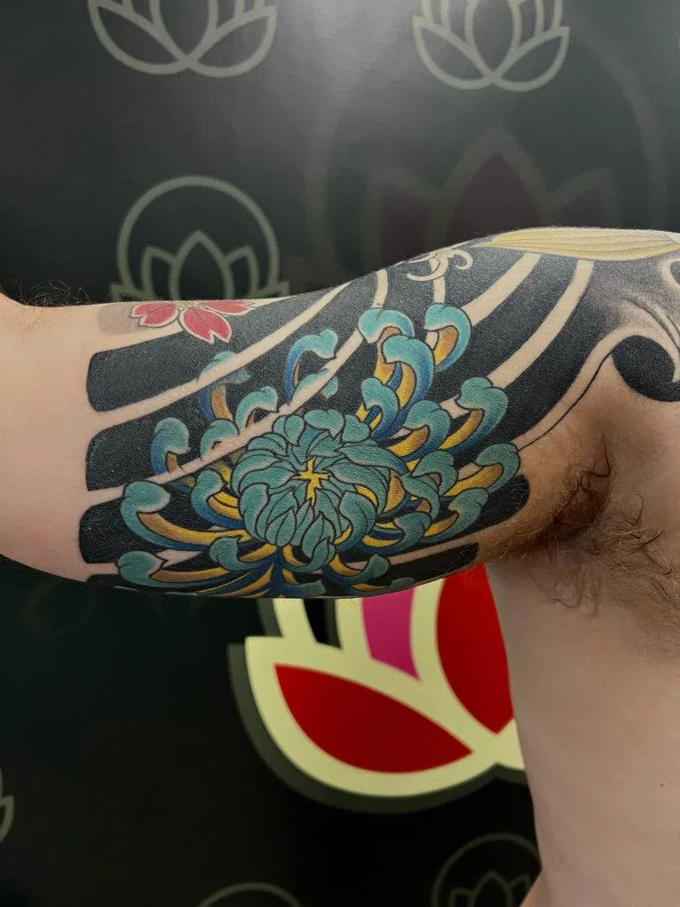 A tattoo on an upper arm featuring a blue and yellow floral design, black stripes, and a small pink flower. The background has faint, stylized lotus patterns.