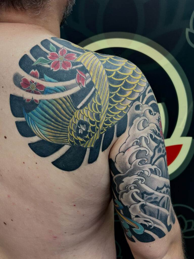 A person's back and upper arm tattoo featuring a colorful koi fish with red flowers on the back and black and grey waves on the upper arm.