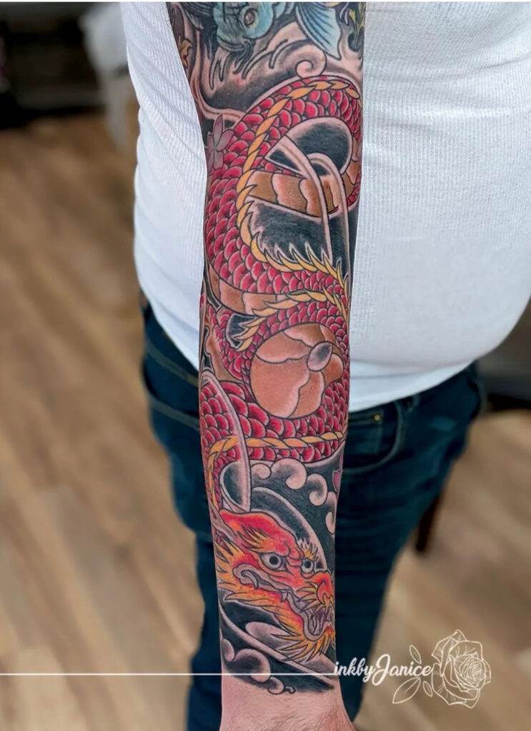 A person with a colorful Japanese-style sleeve tattoo featuring a dragon and koi fish. The person is wearing a white shirt and blue jeans.