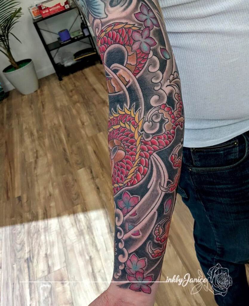 A person with a full sleeve tattoo featuring vibrant colors and intricate designs on their left arm. The background shows a wooden floor, a small shelf, and a house plant.