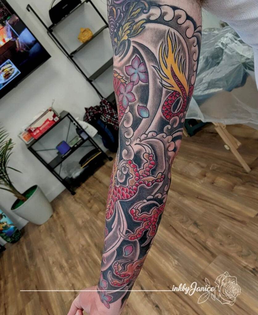 A person's arm is shown with a detailed full-sleeve tattoo featuring colorful dragon and floral designs. In the background, there are shelves with items and a potted plant on a wooden floor.