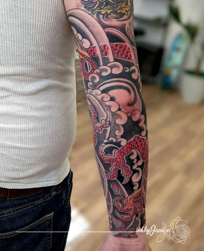 A person with a colorful sleeve tattoo featuring ornate designs and a dragon motif is shown from the back. They are wearing a white sleeveless top and jeans.