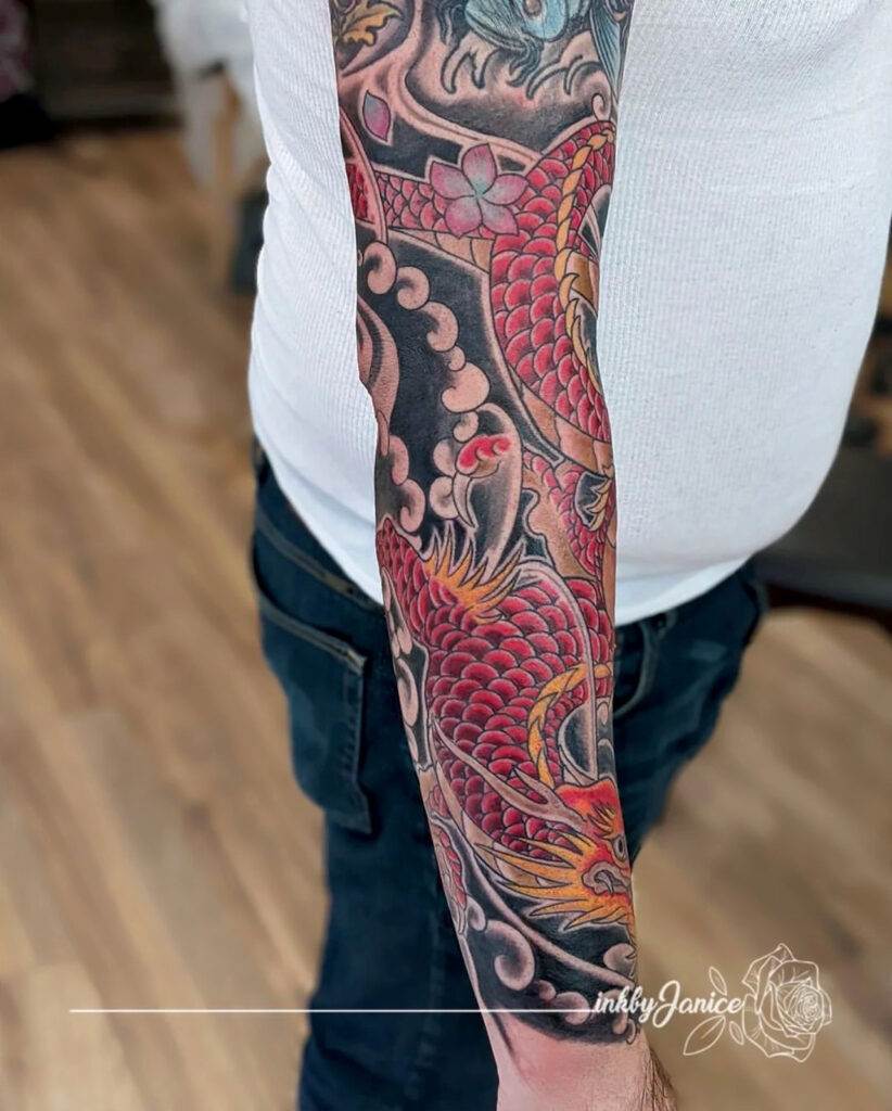 A person with a full sleeve tattoo on the left arm, featuring a detailed red dragon and clouds. The individual is wearing a white sleeveless shirt and jeans.