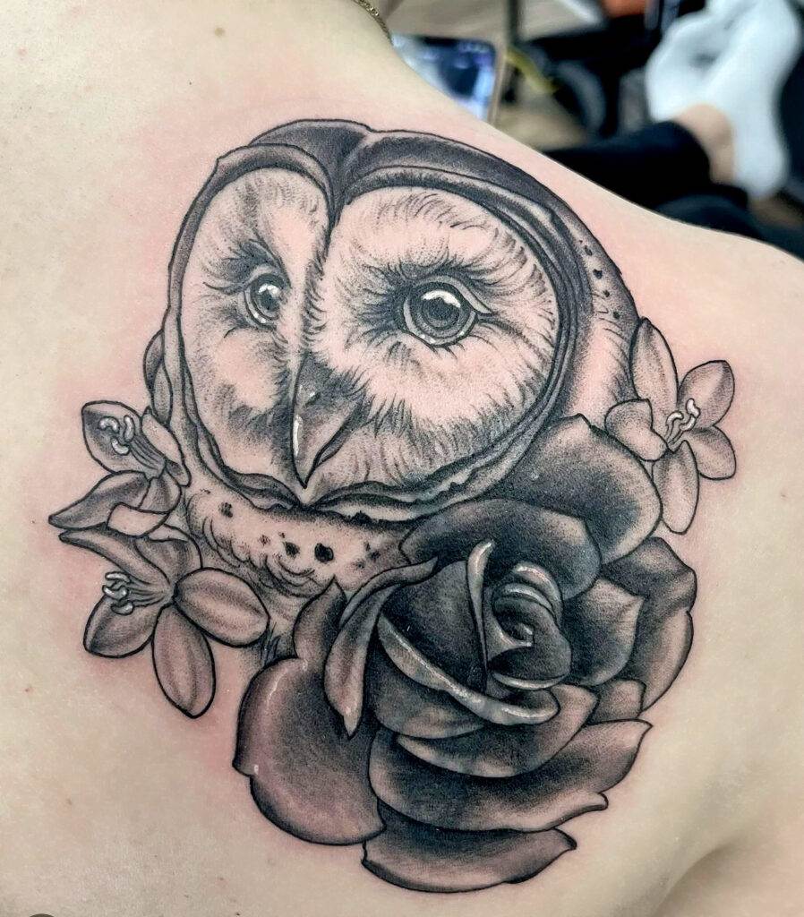 A detailed black and grey tattoo of a barn owl interwoven with roses and leaves on a person's upper back.