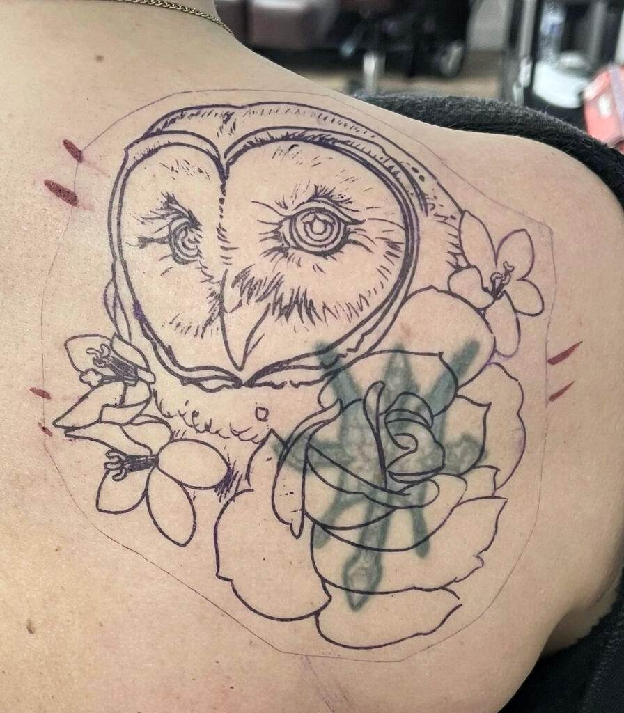 A partially completed tattoo on a person's back, featuring an outline of an owl's face surrounded by flowers.