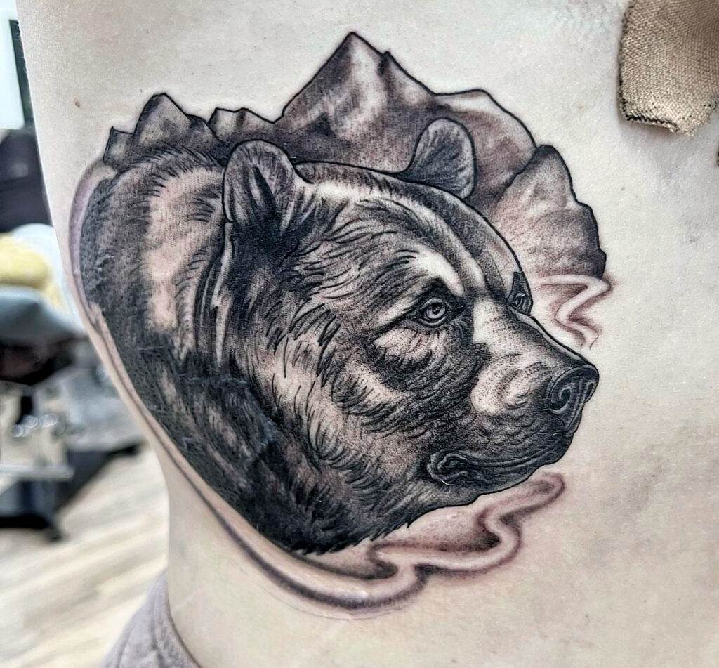 Tattoo of a bear's head with a mountain range background, located on a person's side.