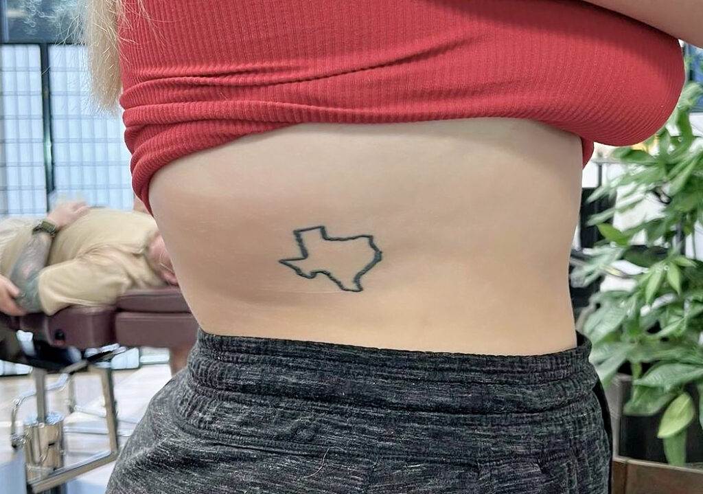 A person with a Texas state outline tattoo on their side is lifting their red shirt to reveal it. Another person is lying on a tattoo chair in the background.