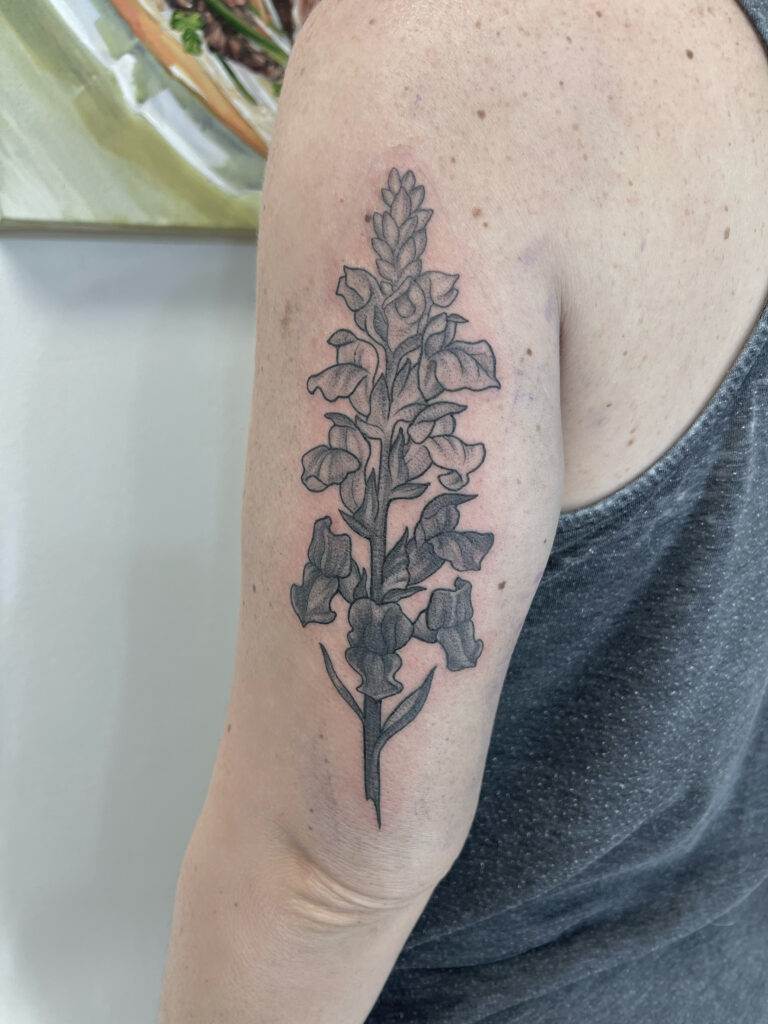 A grayscale floral tattoo on a person's upper arm featuring a detailed and vertical arrangement of flowers and leaves. The person is wearing a sleeveless shirt.