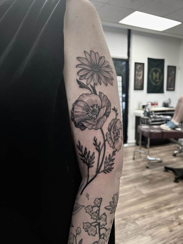 A black and gray flower tattoo featuring poppies and a daisy is on a person's upper arm in a tattoo studio.