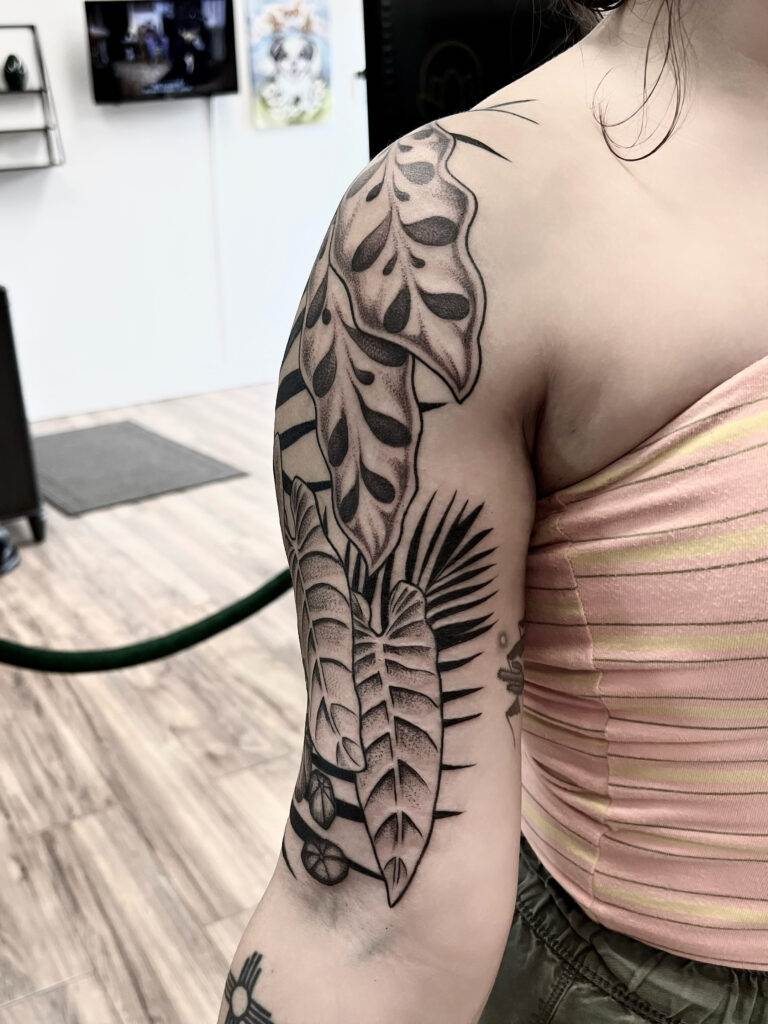 A person with a full sleeve tattoo on their right arm featuring large tropical leaves and intricate shading, wearing a pink striped top.