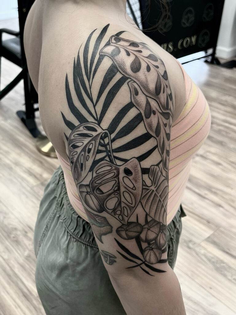 A person with a detailed black and gray tropical plant tattoo covering their shoulder and upper arm. The person is wearing a yellow striped tank top and green pants.