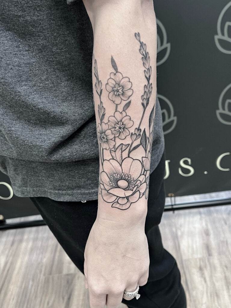 A forearm tattoo featuring a floral design with various flowers in black ink, displayed on an individual wearing a gray shirt and black pants.