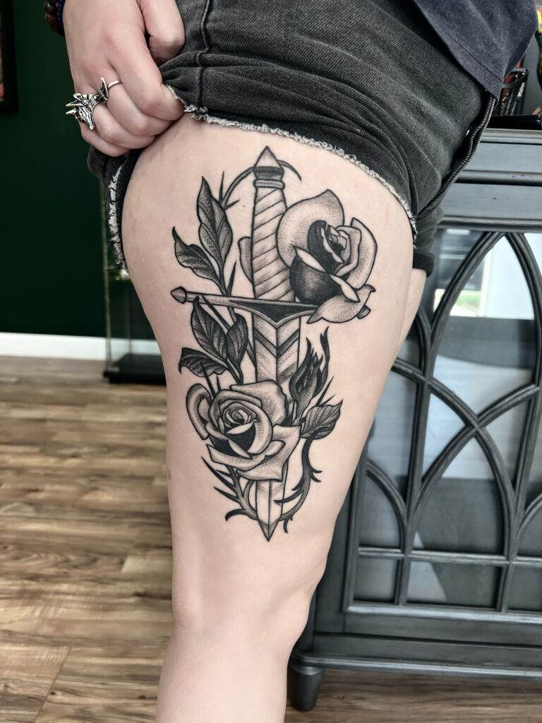 Thigh tattoo of a sword entwined with roses, shown on a person wearing black shorts.