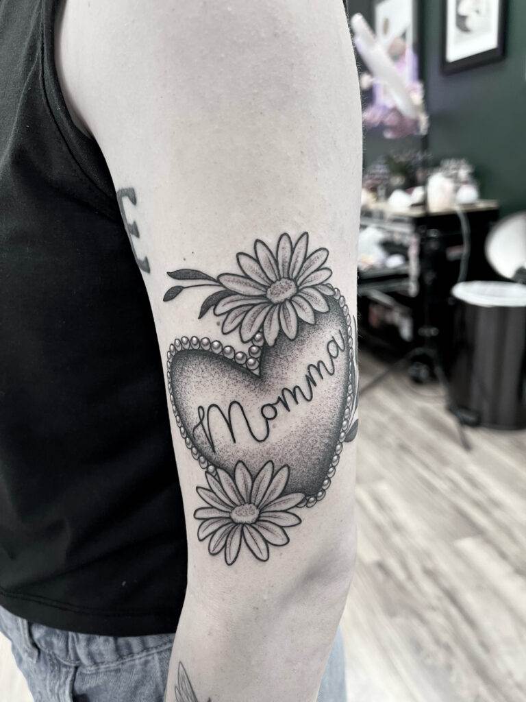 A black and white tattoo of a heart with the word "Momma" inside, surrounded by daisies, on a person's upper arm.