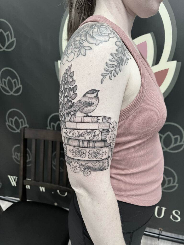 A person with an upper arm tattoo featuring a bird perched on stacked books with floral designs around it. The individual is wearing a pink sleeveless top.