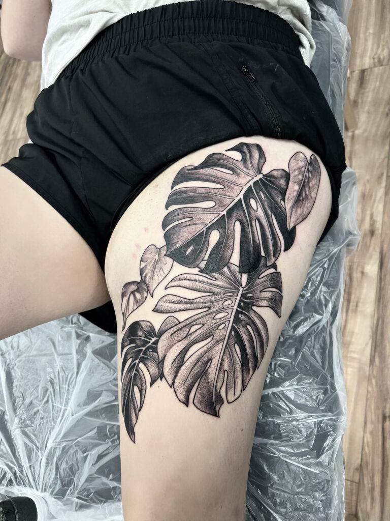 A person with a detailed black and grey tattoo of Monstera leaves on their upper thigh, wearing black shorts, lying down during the tattoo process.