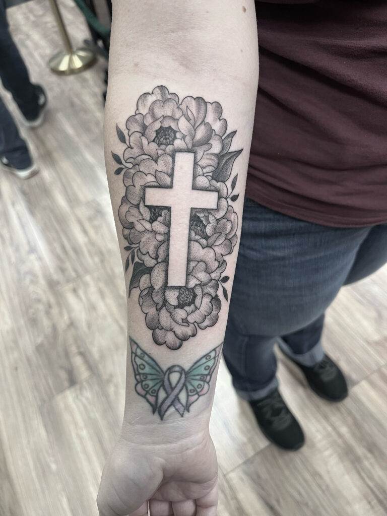 Forearm tattoo featuring a cross surrounded by flowers and a butterfly with a ribbon below it.