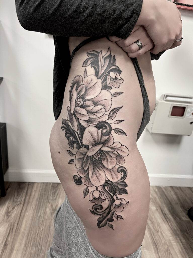 A person displays a detailed black and grey floral tattoo covering their upper thigh and hip.