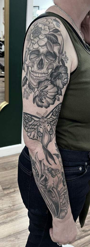 A person with a detailed black and grey tattoo sleeve featuring a skull, flowers, and a moth on their left arm.