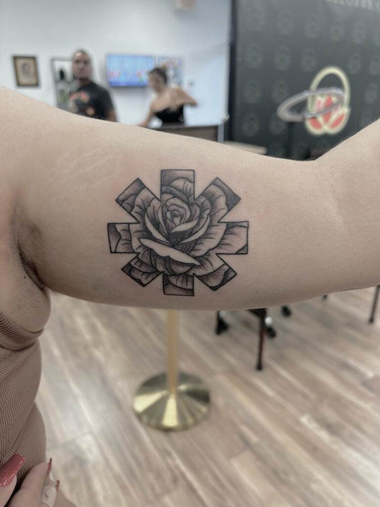 A person displays a tattoo on their bicep featuring a detailed rose design within a geometric star shape. Other people are slightly visible in the background in a room with wooden flooring.