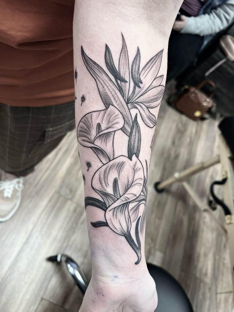 A tattoo of blooming calla lilies is inked on a person's inner forearm. The tattoo is detailed with shading and linework, showcasing multiple flowers and leaves.