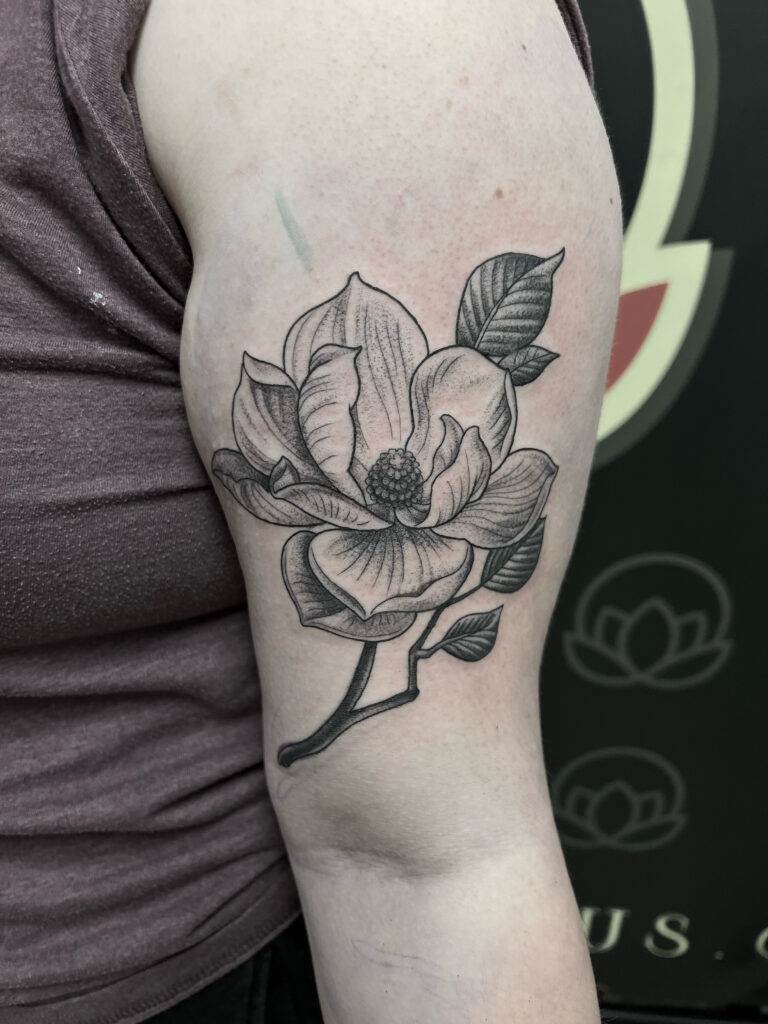 A detailed, black and gray magnolia flower tattoo is on the upper arm of a person wearing a maroon sleeveless shirt.