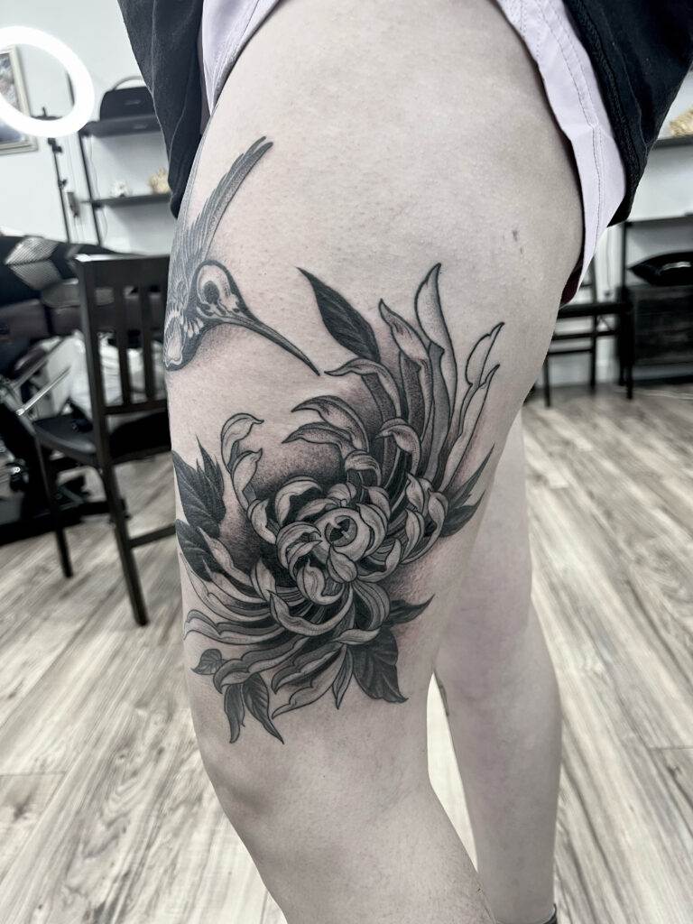 A black and gray tattoo on the upper thigh featuring a hummingbird hovering near a large chrysanthemum flower.