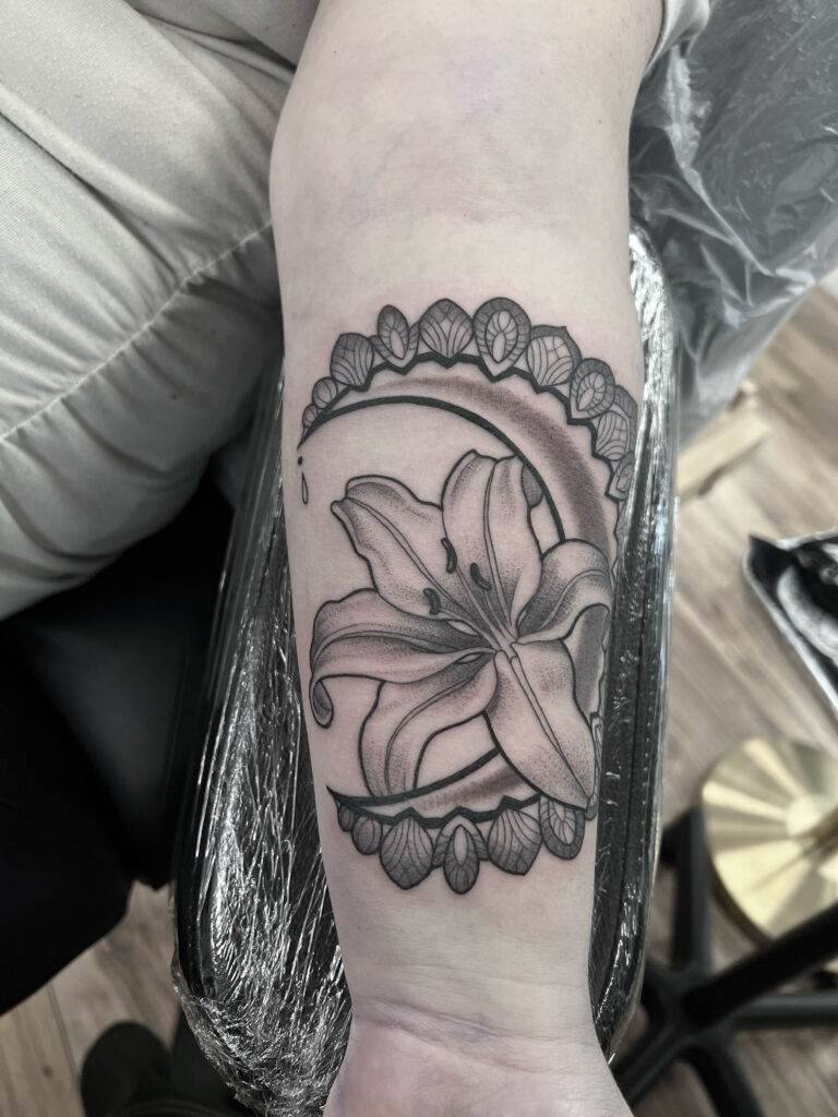 A black and grey tattoo of a lily flower within a circular design decorated with leaf-like patterns on a person's forearm.