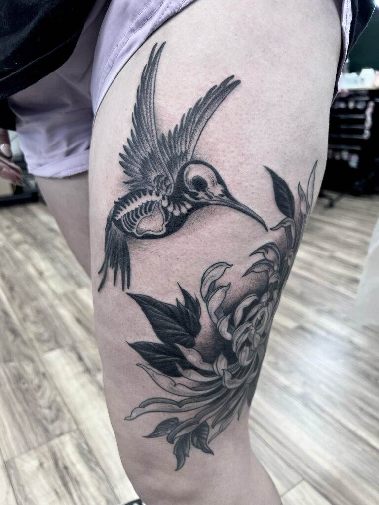 A black and gray tattoo of a hummingbird with visible skeleton details and a floral design on a person's upper thigh.