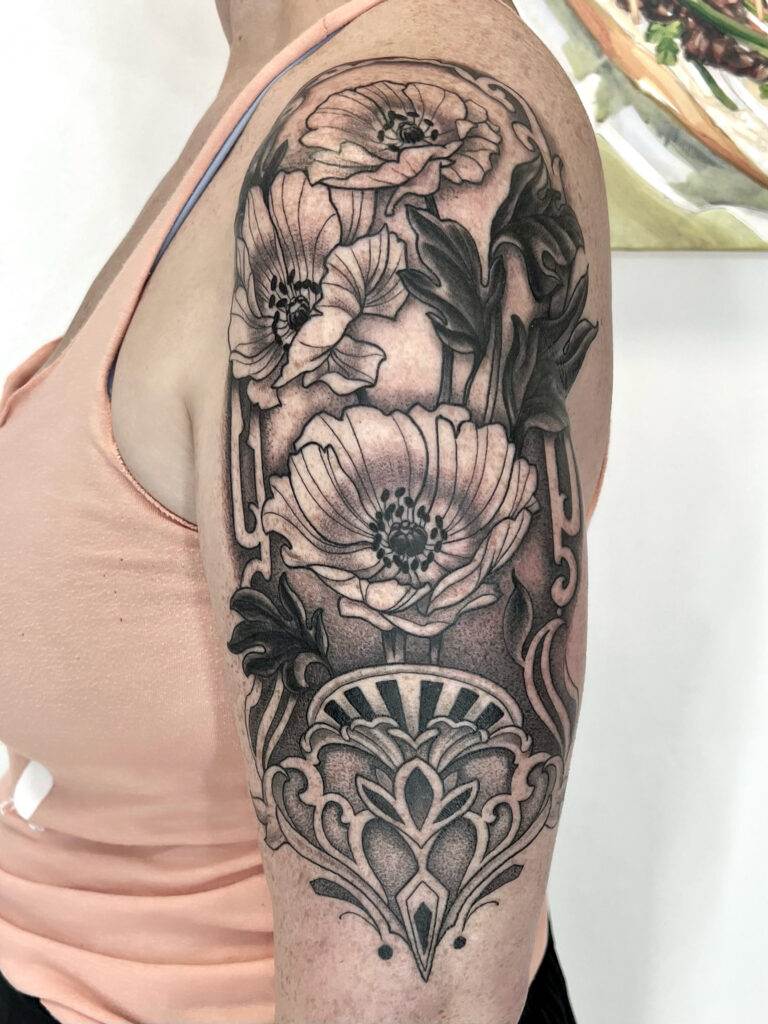 A person with a floral tattoo design on their upper arm, featuring detailed flowers and ornamental patterns in black and gray ink. They are wearing a sleeveless pink top.