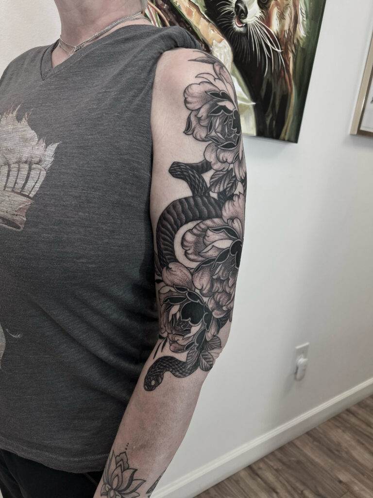 A person with a detailed black and gray sleeve tattoo featuring flowers and a snake, wearing a sleeveless gray shirt.