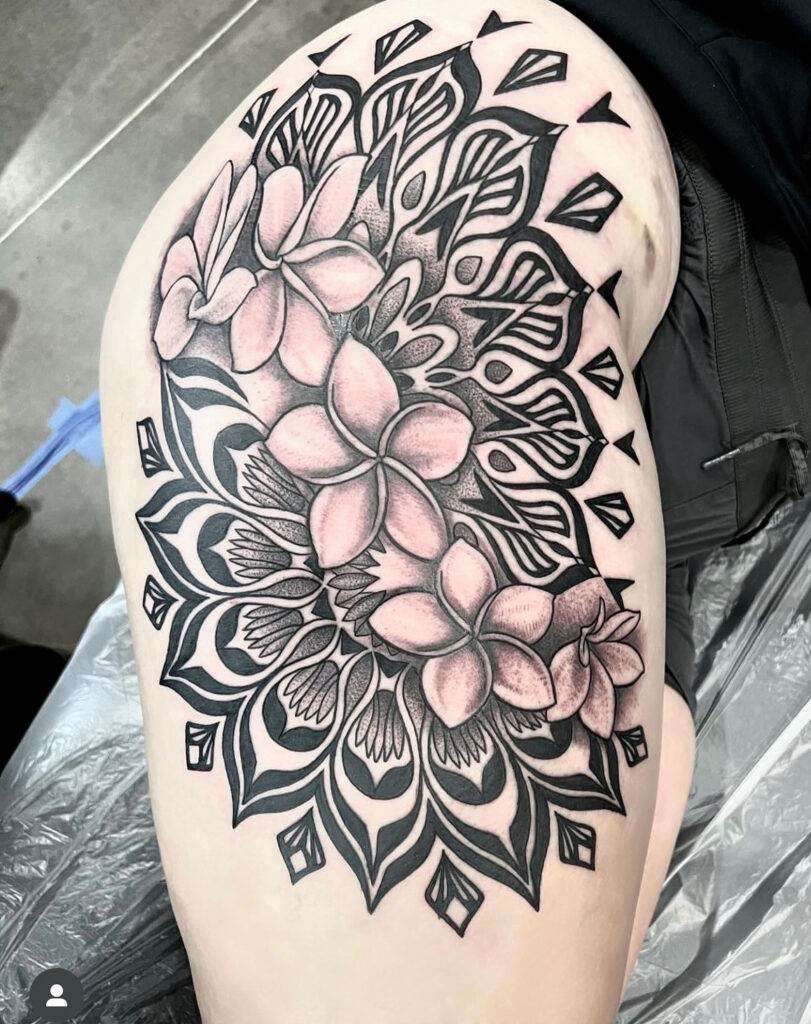 A person's thigh with a detailed black and grey tattoo of flowers and geometric patterns.