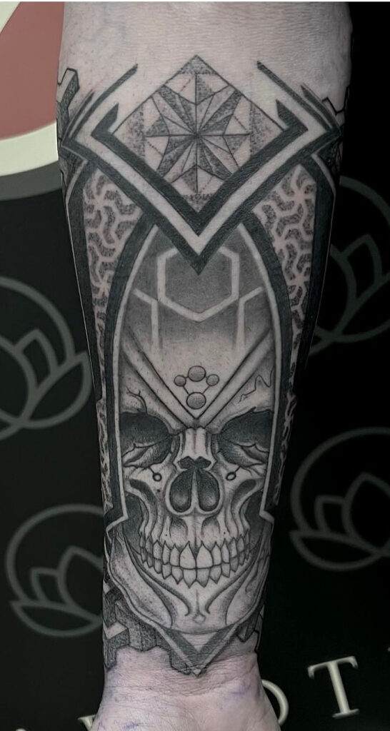 A black and grey tattoo on a forearm depicts an intricately detailed skull with geometric patterns and symmetric shapes above and around it.