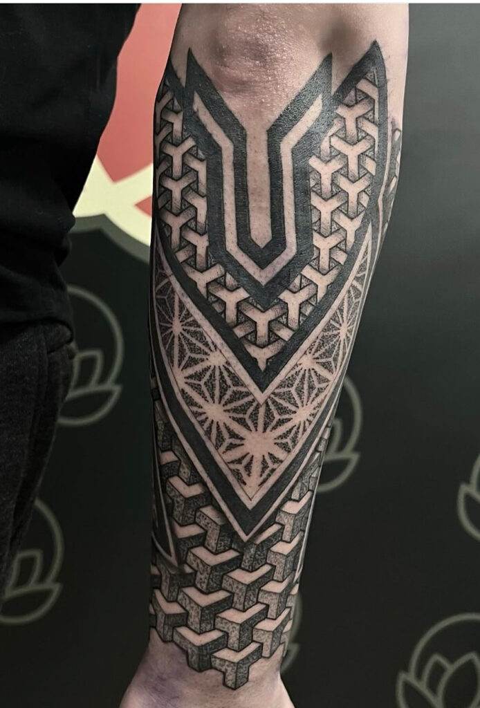 A heavily tattooed forearm featuring geometric patterns and interlocking designs, extending from elbow to wrist.