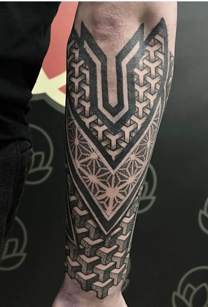 A forearm displaying an intricate geometric black tattoo with various patterns and shapes, set against a dark background.