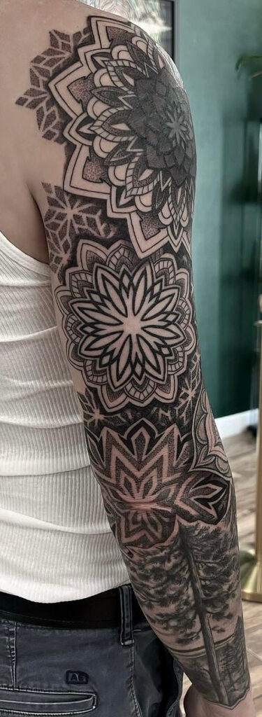 A person's arm with an intricate geometric and floral tattoo sleeve in black and grey ink. The person is wearing a white sleeveless shirt and blue jeans. The background is a green wall and wooden floor.