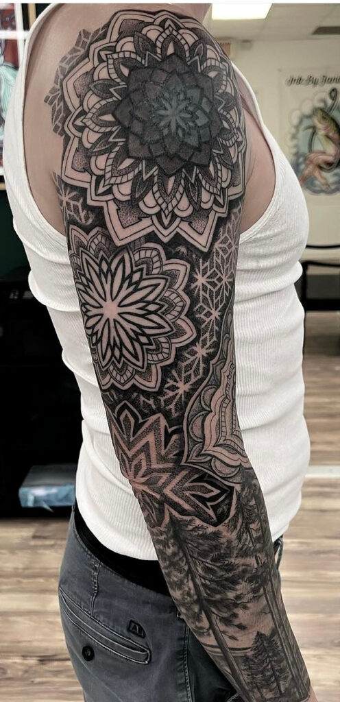 A person with a full sleeve tattoo featuring intricate geometric and floral patterns is shown from the side, wearing a white sleeveless shirt and grey pants.