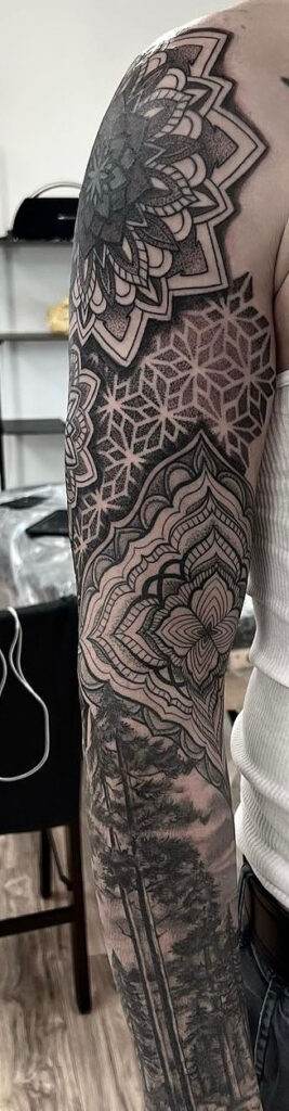 A close-up of a person's upper arm covered in intricate black and grey tattoos, featuring geometric shapes and mandalas, extending down to the forearm with tree designs.