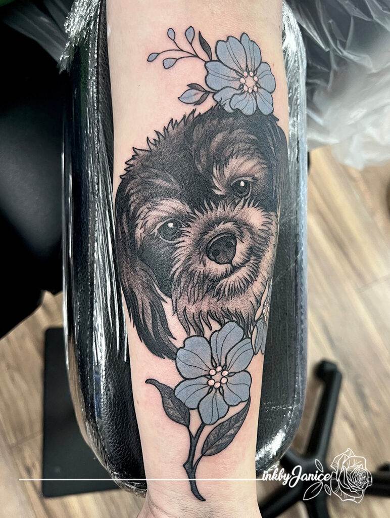 A tattoo of a dog's face with floral decorations on a person's forearm, featuring purple flowers and green leaves.