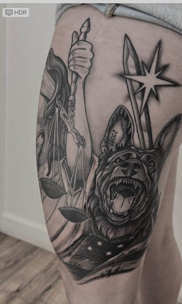 A thigh tattoo featuring Lady Justice holding scales and a sword, alongside a snarling dog with an American flag.