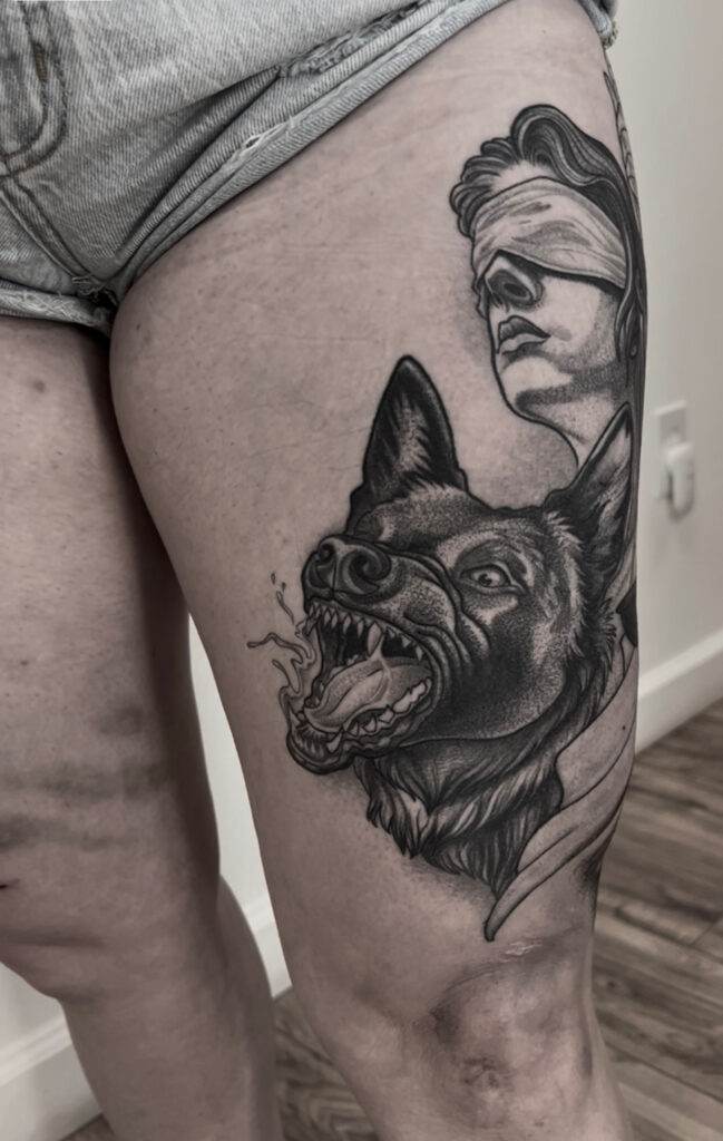 Black and grey tattoo on a person's thigh, depicting a blindfolded figure and a growling wolf.