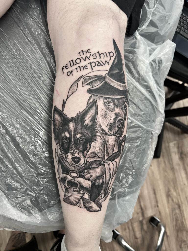 A black and gray tattoo on a person's leg depicts two dogs dressed as characters from "The Lord of the Rings," with the words "The Fellowship of the Paw" above them.