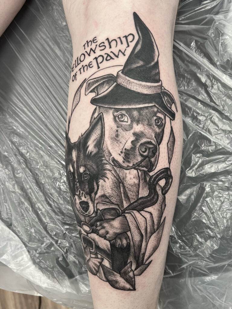 A tattoo on a person's leg depicting two dogs from "The Fellowship of the Paw," one wearing a wizard hat and the other resembling a wolf, with detailed shading and line work.