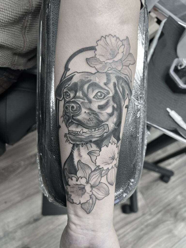 Black and white tattoo of a dog's face surrounded by large flowers on a person's forearm.